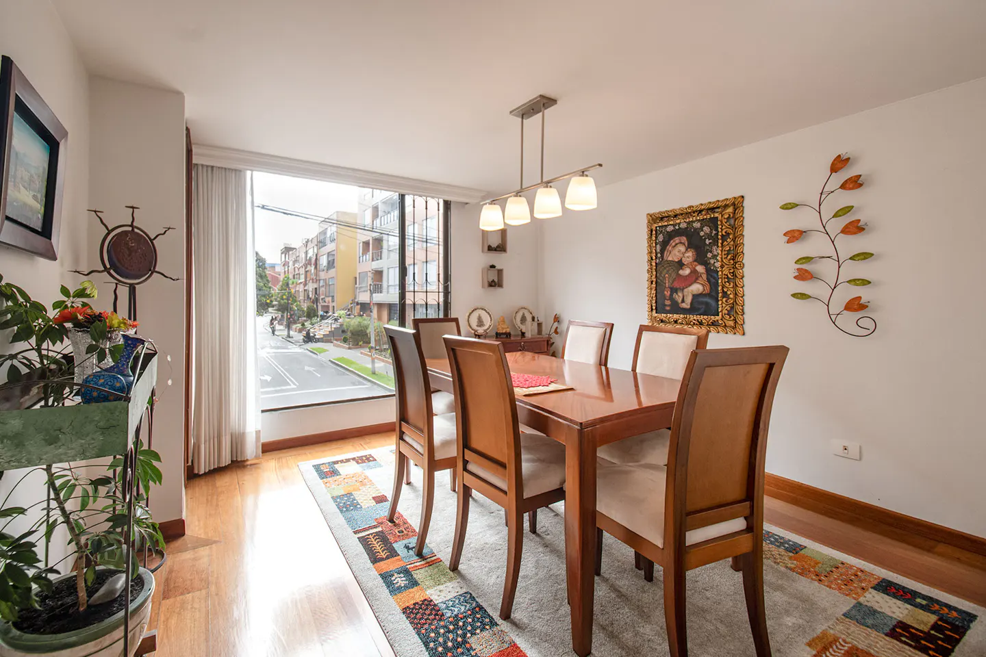 Dining room with a wooden table, six chairs, a colorful patterned rug, and a large window overlooking a city street.