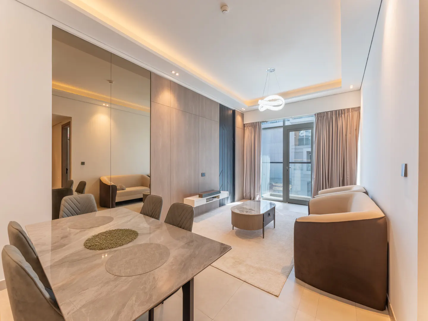 A modern living room with a marble table, gray chairs, and a beige sofa. A large mirror reflects the room, enhancing the sense of space.