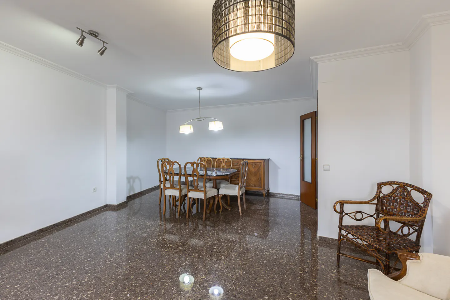 Bright dining room with a table, chairs, and a wooden cabinet on a speckled floor. A wicker chair sits near a white sofa.