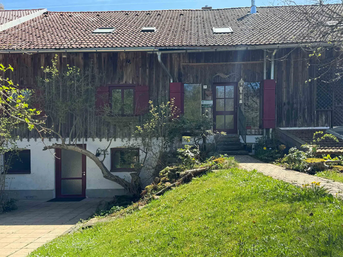 Exterior view of a two-story house with a brown wood facade, red shutters, and a red tile roof. A green lawn slopes up to the house.