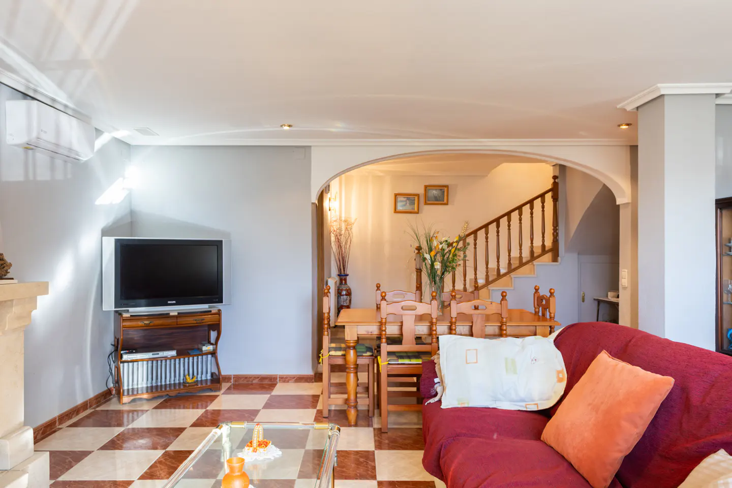 Living room with red sofa, glass coffee table, TV on a wooden stand, and a dining area with wooden chairs and staircase.