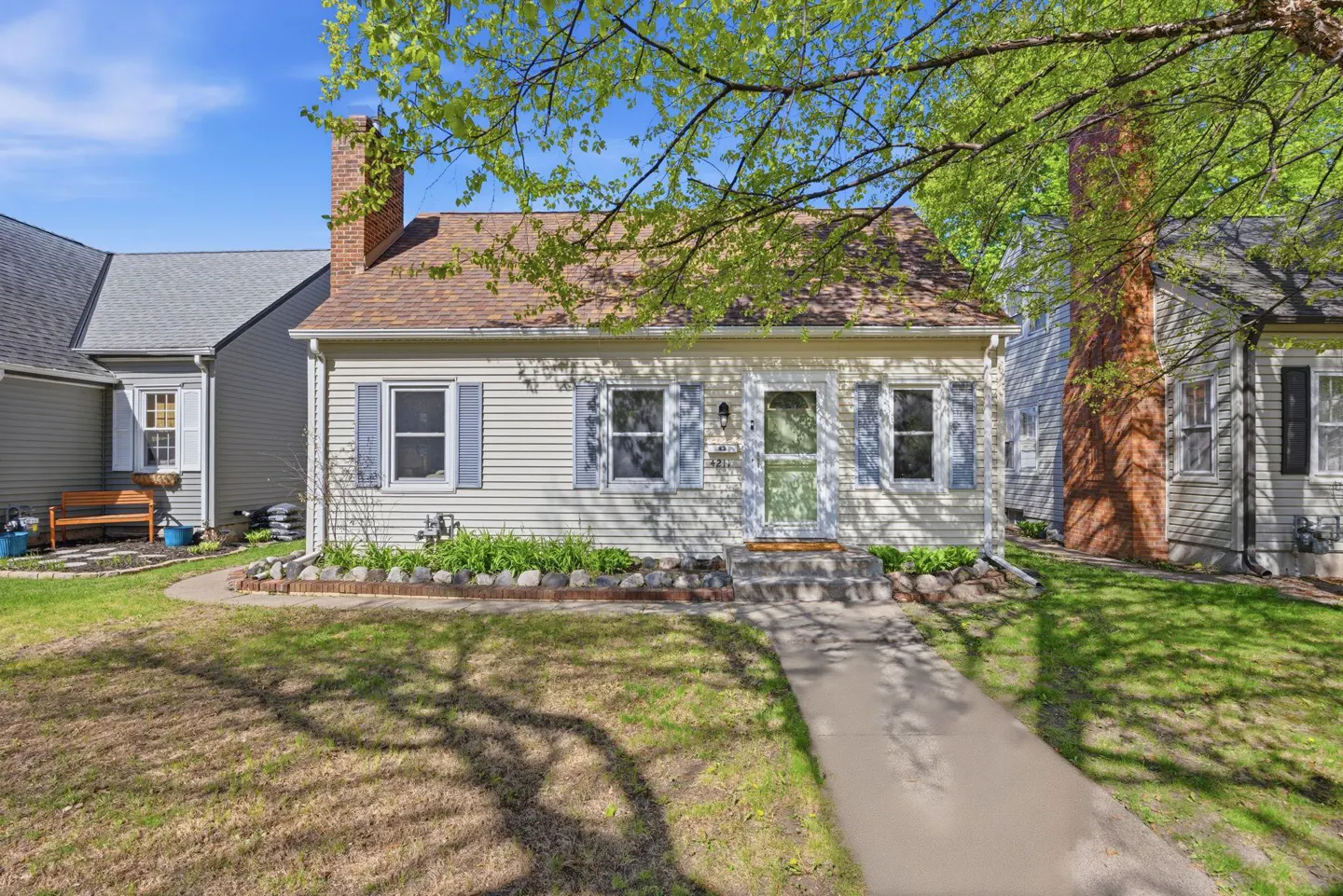 A cream-colored house with blue shutters, a brown roof, and a brick chimney on a sunny day.