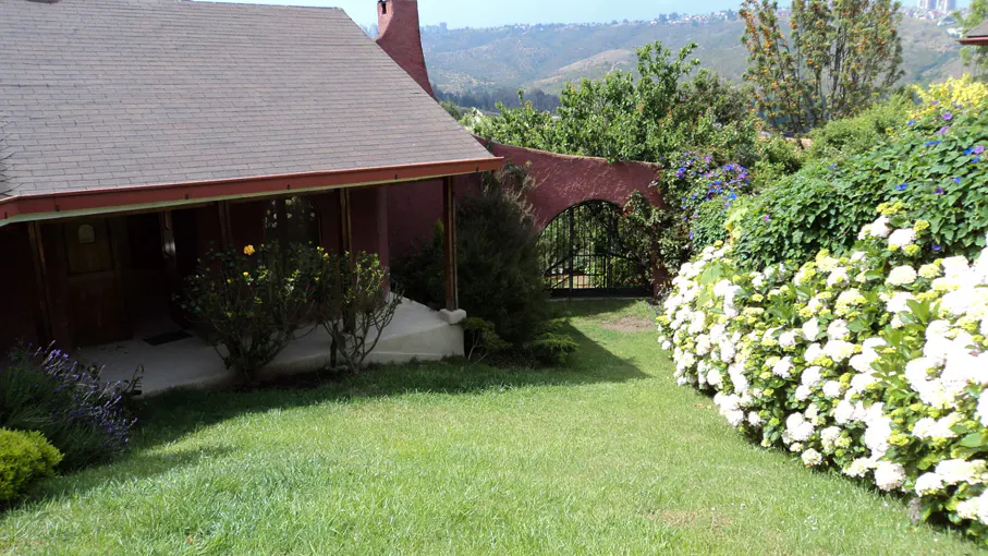 Exterior view of a red house with a brown roof, green lawn, and white hydrangeas. A black gate is visible in the background.