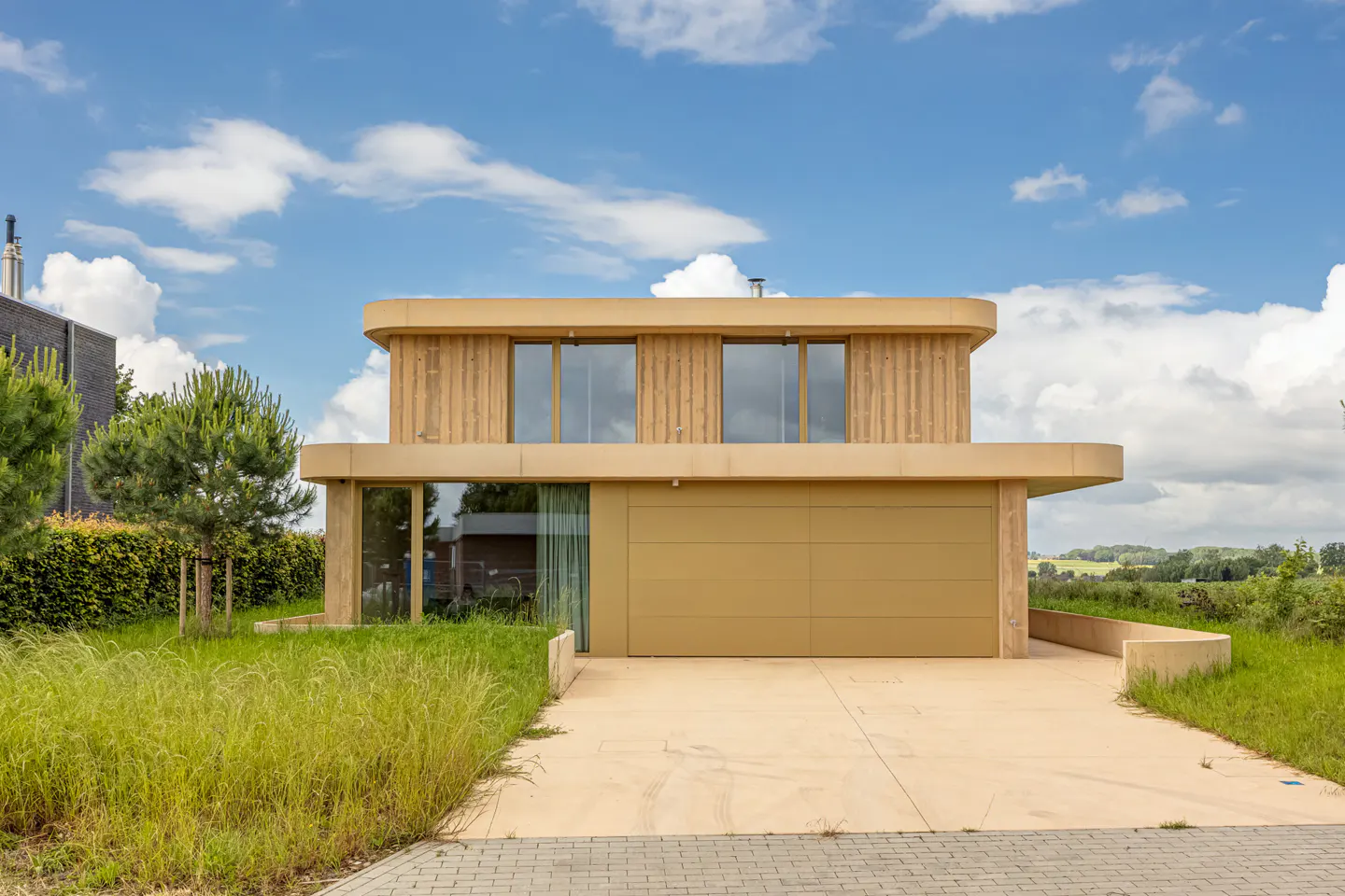 Modern two-story house with light wood siding, large windows, and a beige garage door under a blue sky with white clouds.