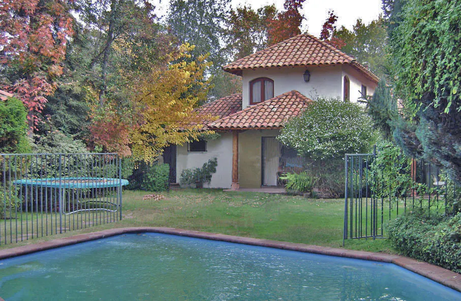 A white house with a red tile roof is surrounded by trees and a blue swimming pool.
