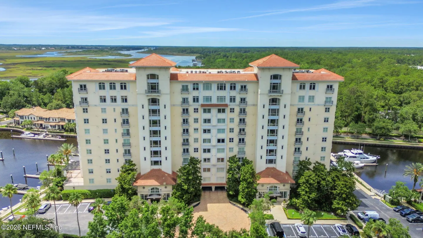 Aerial view of a tall, light-yellow condo building with a red tile roof, surrounded by trees, a river, and a parking lot.