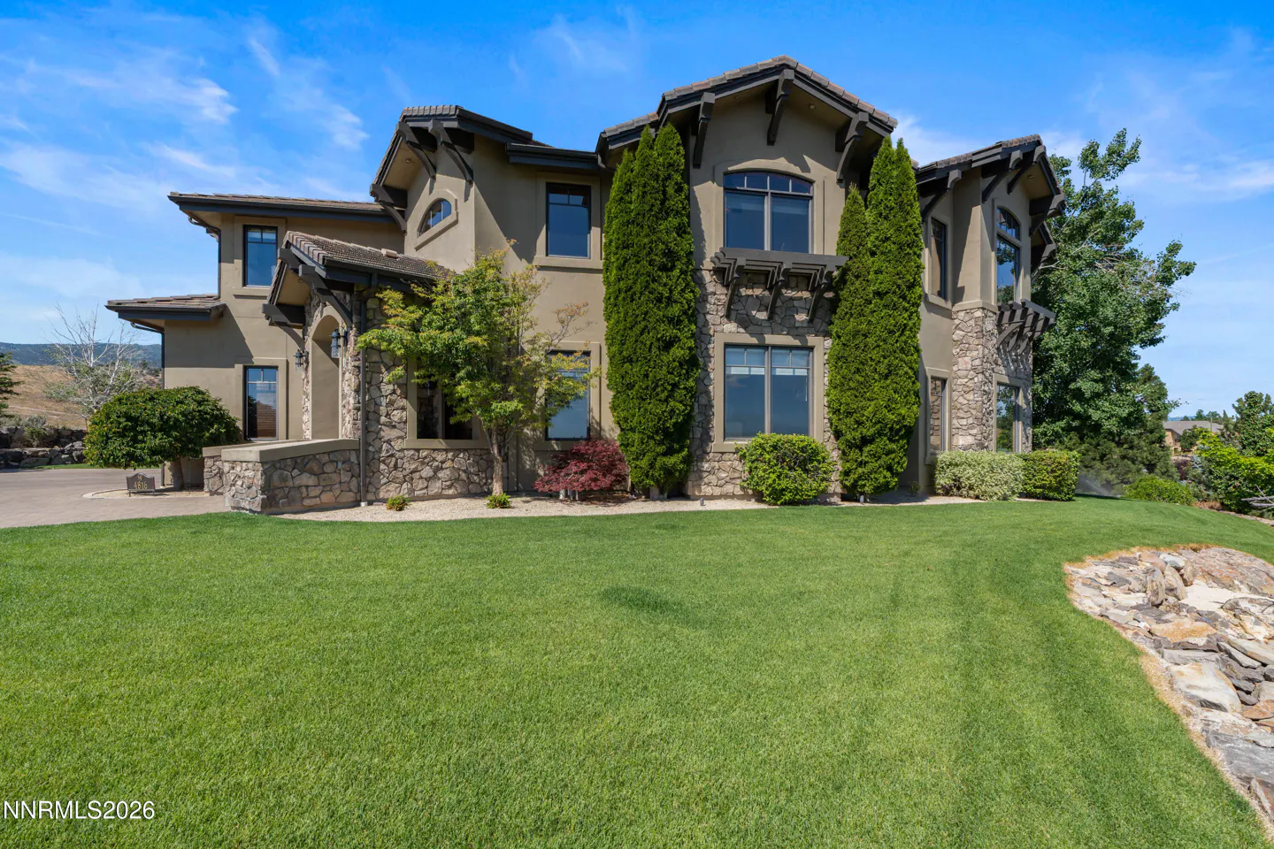 Large two-story stucco and stone luxury home with a manicured green lawn, tall cypress trees, and a bright blue sky.