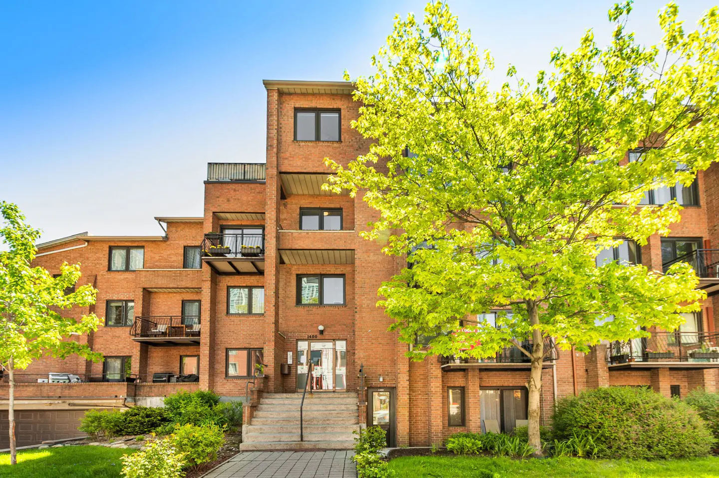 Exterior view of a brick apartment building with balconies, green trees, and a blue sky.