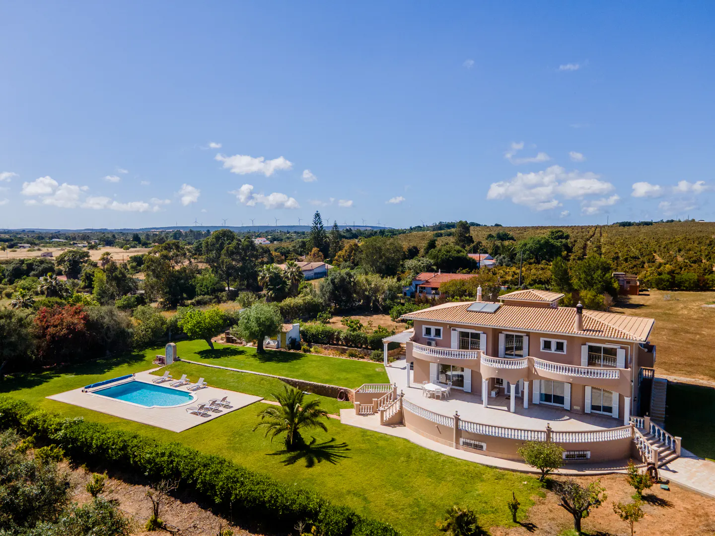 Aerial view of a tan two-story house with a pool, green lawn, and trees under a blue sky.