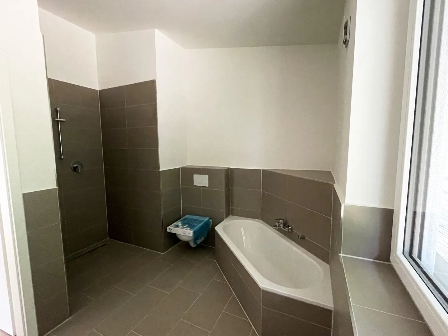 A bathroom with gray tile, a white bathtub, and a toilet with a blue cover.