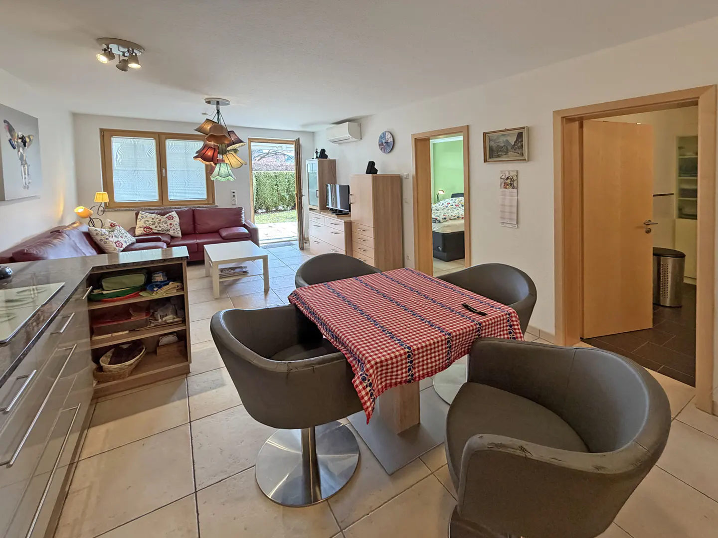 Open-concept living space with a dining table covered in a red and white checkered cloth, a red sofa, and an open door to a green yard.