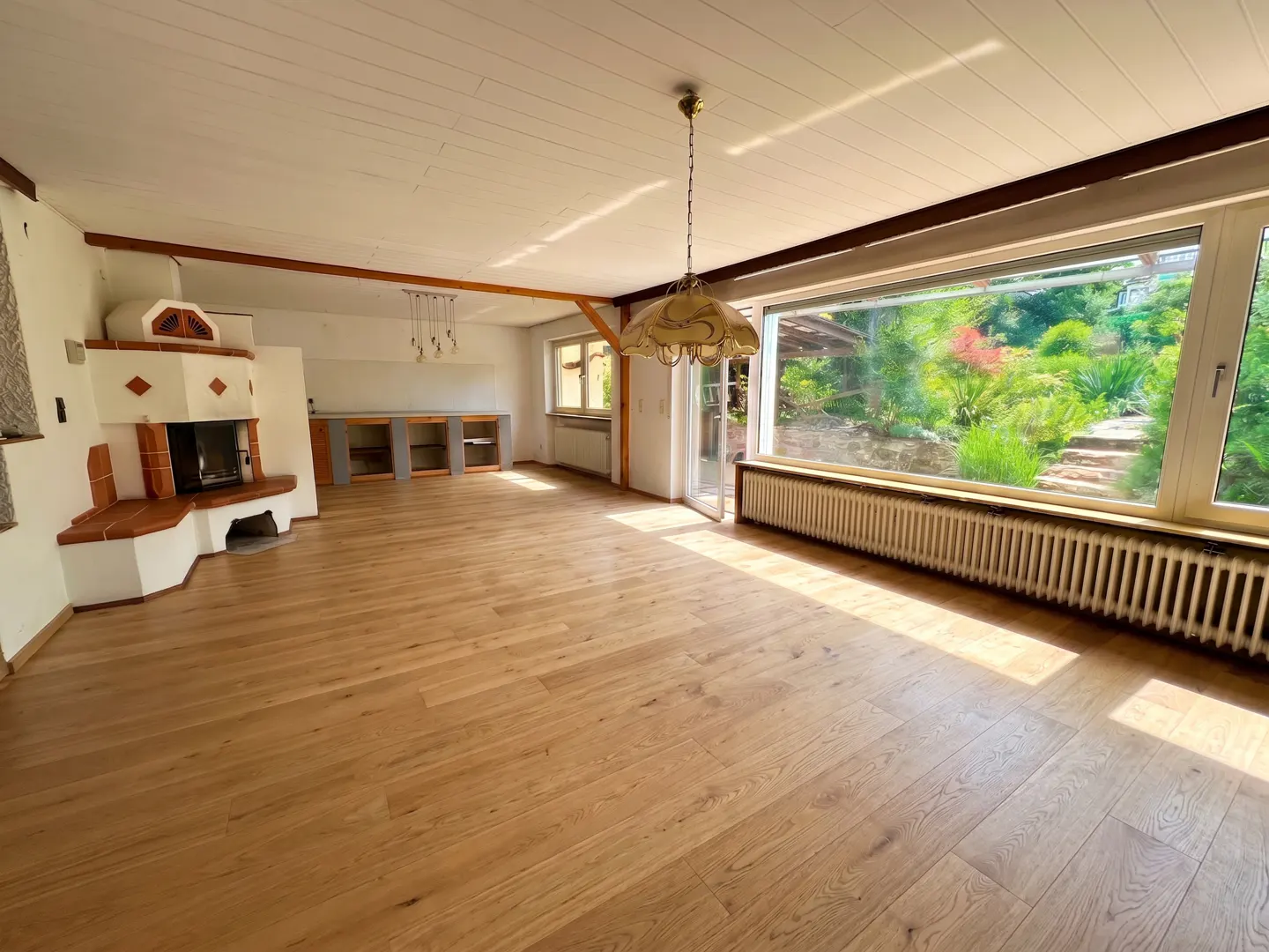 Bright, empty living room with wood floors, a fireplace, and a large window overlooking a lush garden.