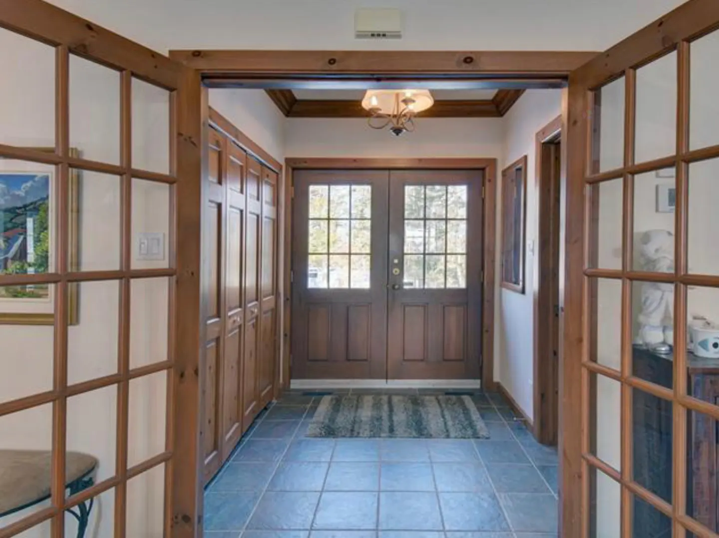 A home's entryway features double doors, wood trim, blue tile flooring, and a small chandelier.