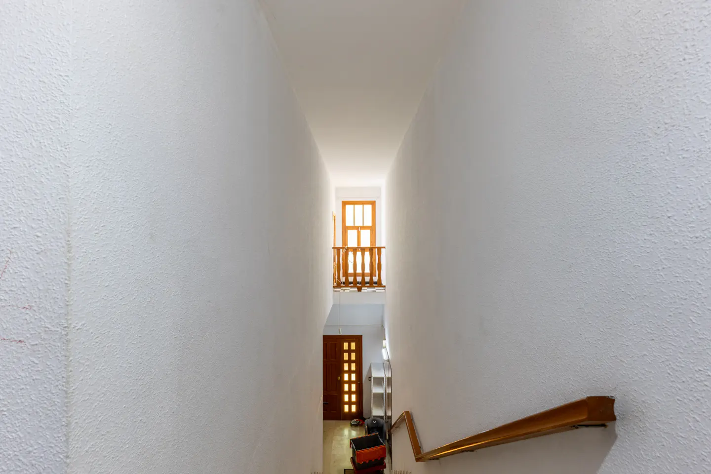 Hallway view from stairs with white walls, a wooden handrail, and a door and window at the end.