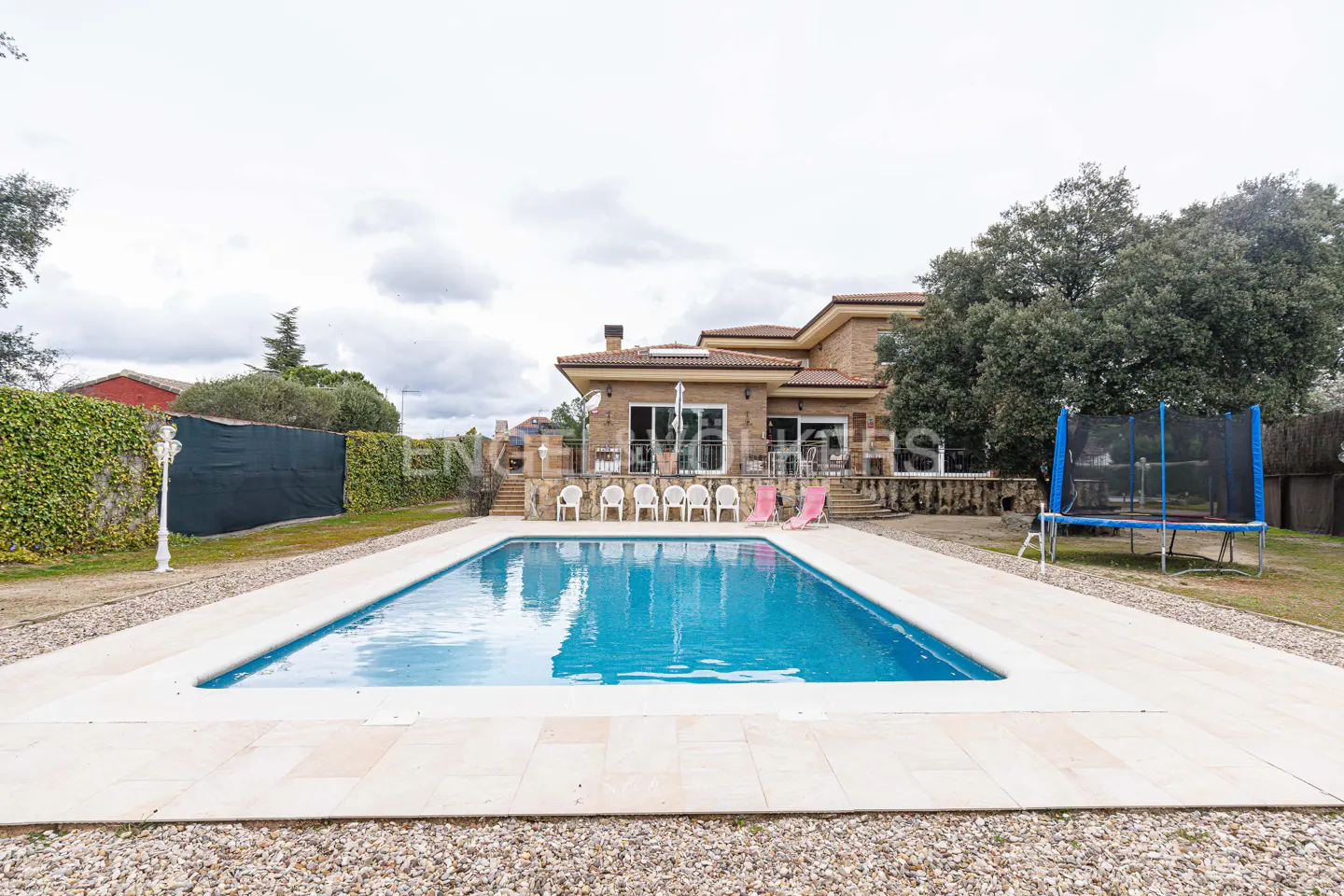 A backyard with a pool, chairs, and a trampoline. The pool is surrounded by light-colored tiles and small rocks. A brick house is in the background.