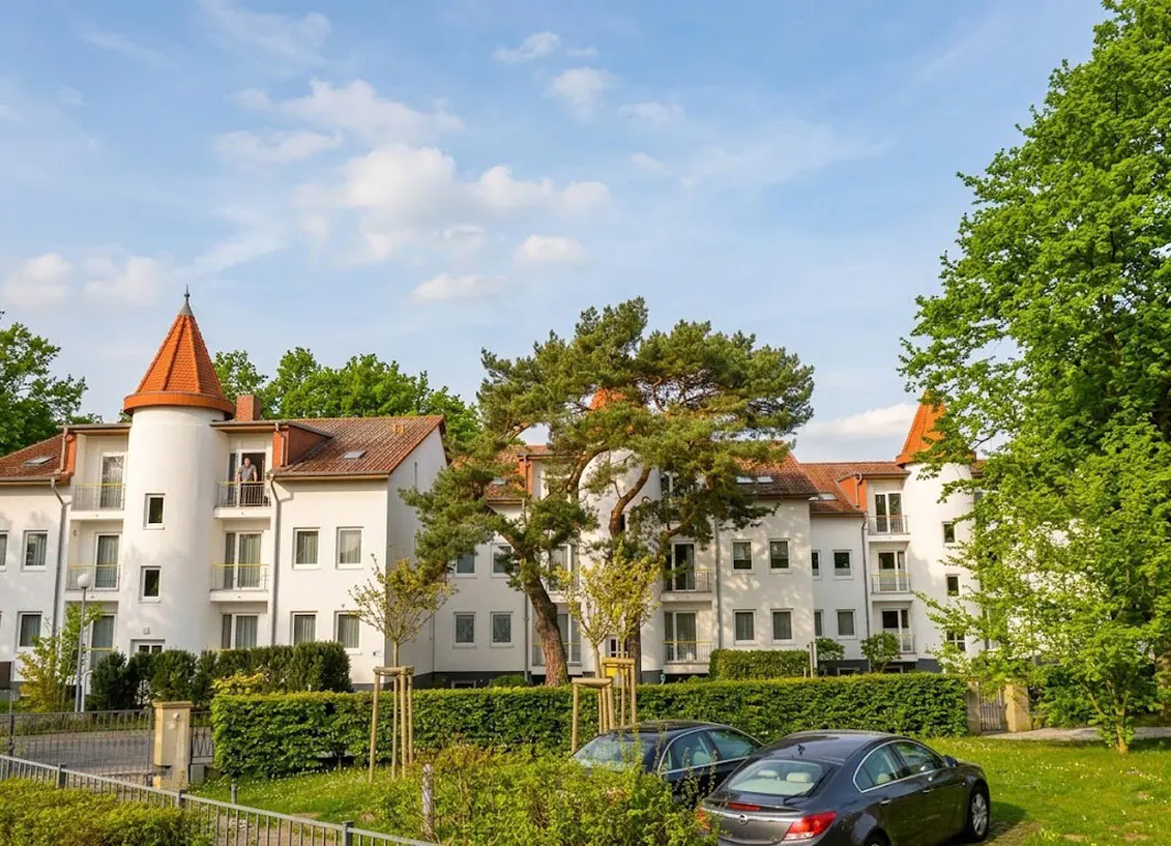 Exterior view of a white apartment building with a red-tiled roof and turret, surrounded by green trees and parked cars.
