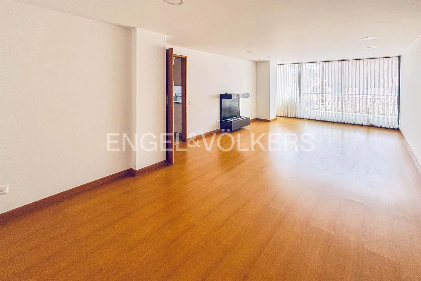 A bright, empty living room with hardwood floors, white walls, and a modern fireplace. A large window with white blinds lets in natural light.