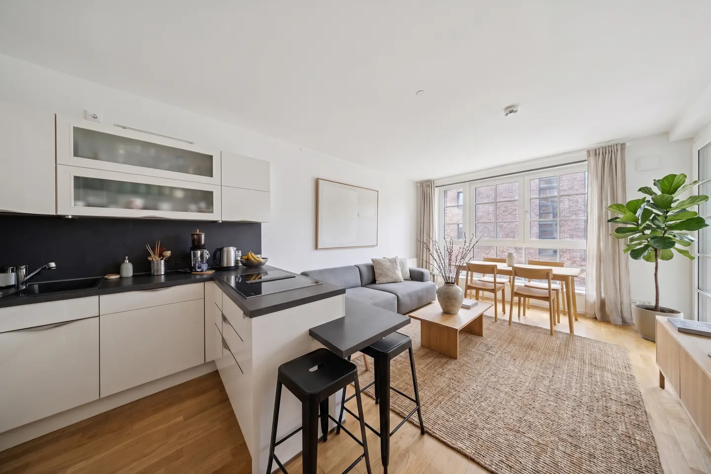 Bright, modern apartment interior with white kitchen cabinets, black countertops, gray sofa, wood dining table, and large windows.