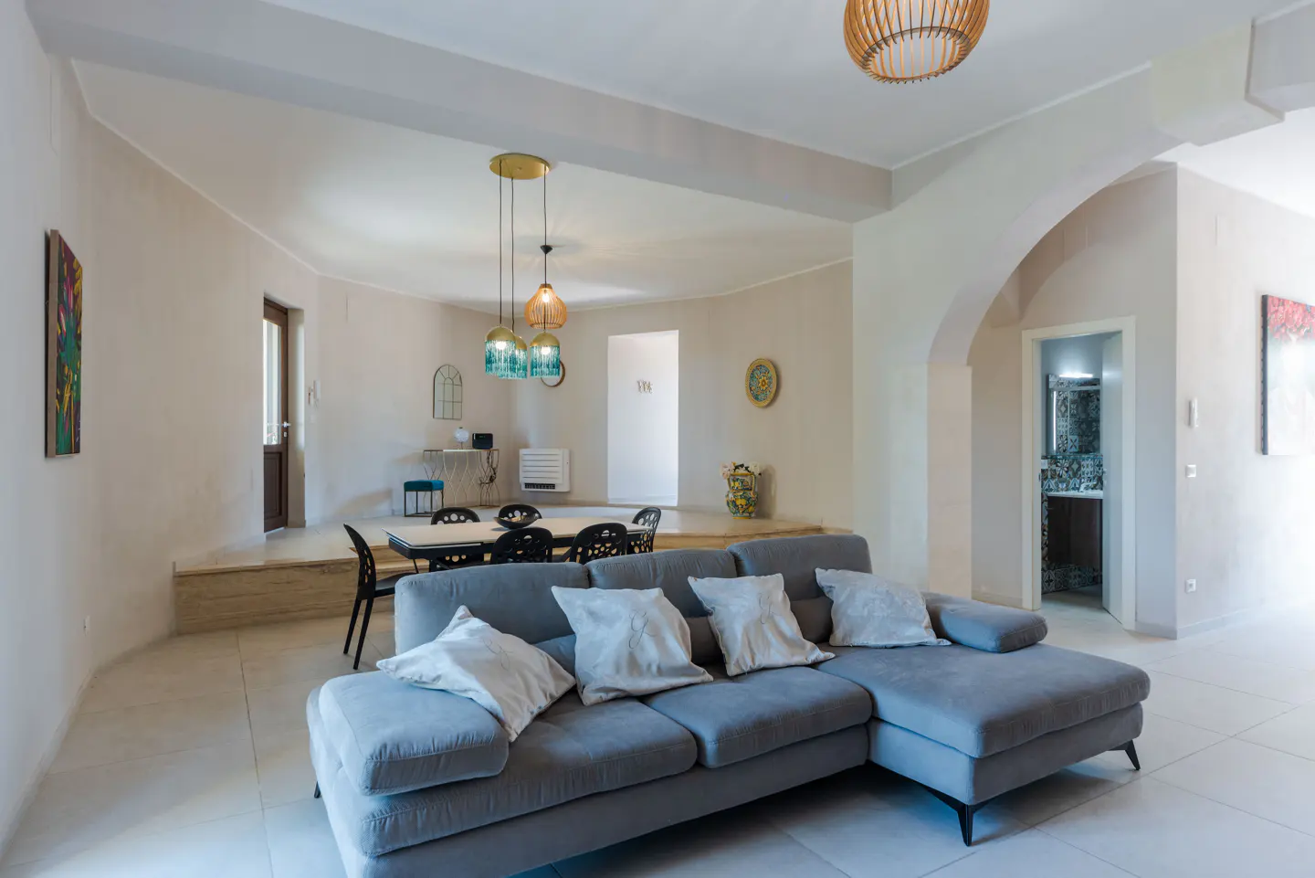 Open-concept living space with a gray sectional sofa, dining table, and pendant lights. Neutral walls and tile floors.