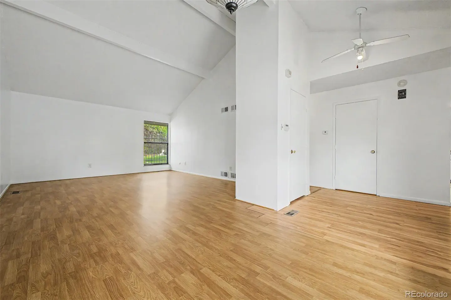 Bright, empty room with light wood floors, white walls, and a vaulted ceiling. A window and two doors are visible.