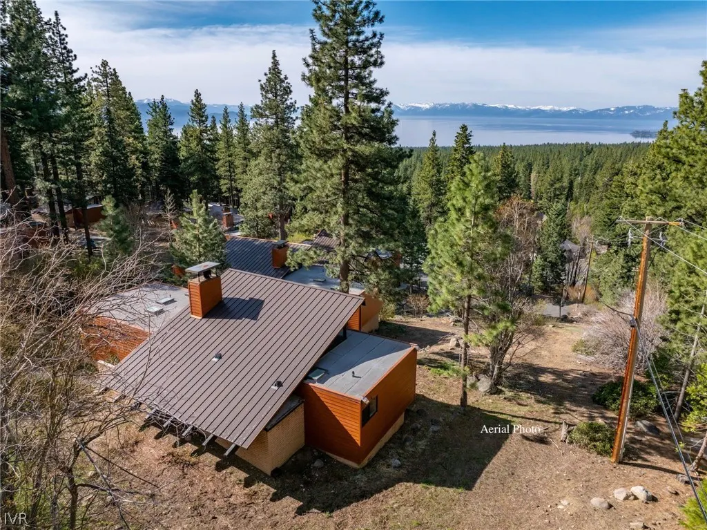 Aerial view of a brown roofed house surrounded by tall green trees, with a lake and mountains in the background.