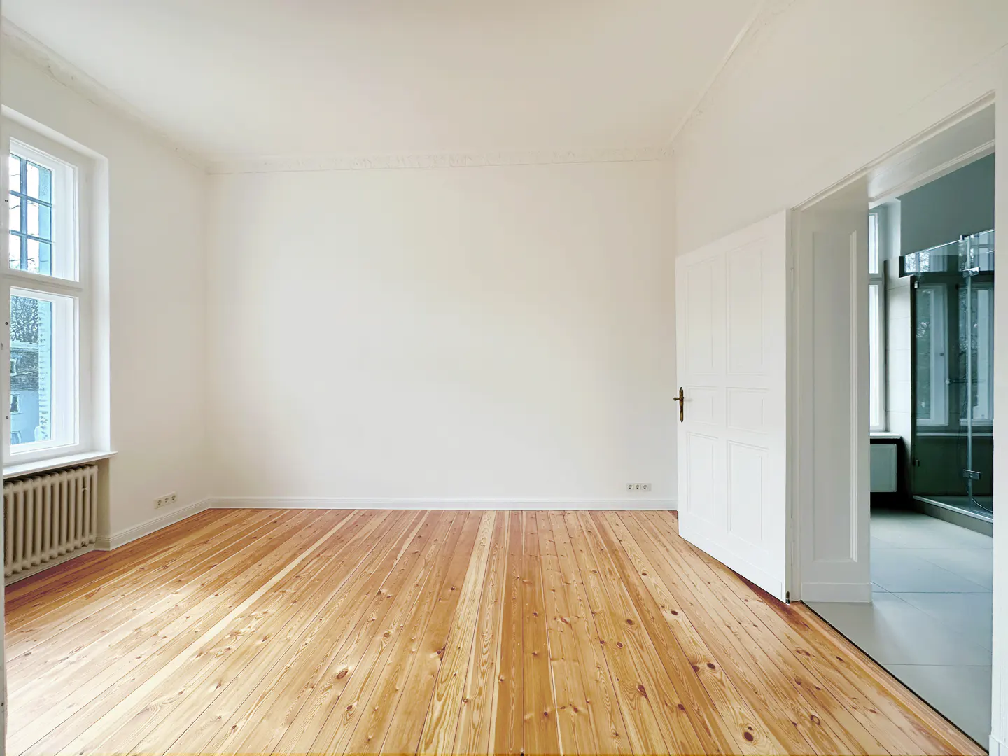 Bright, empty room with light wood floors, white walls, and a window. An open door reveals a glimpse of another room.