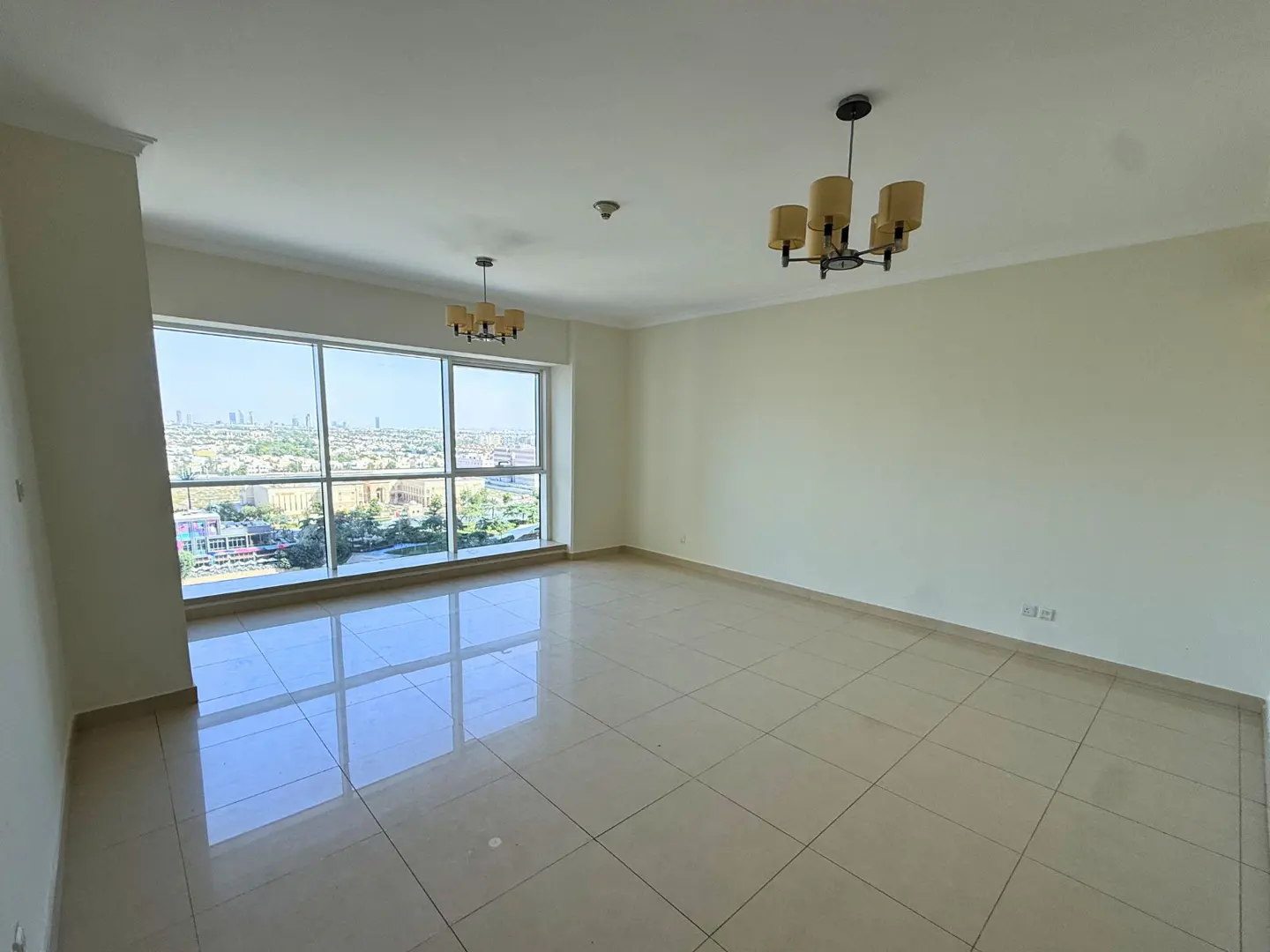 An empty, bright room with beige tile floors, white walls, and a large window with a city view. Two chandeliers hang from the ceiling.