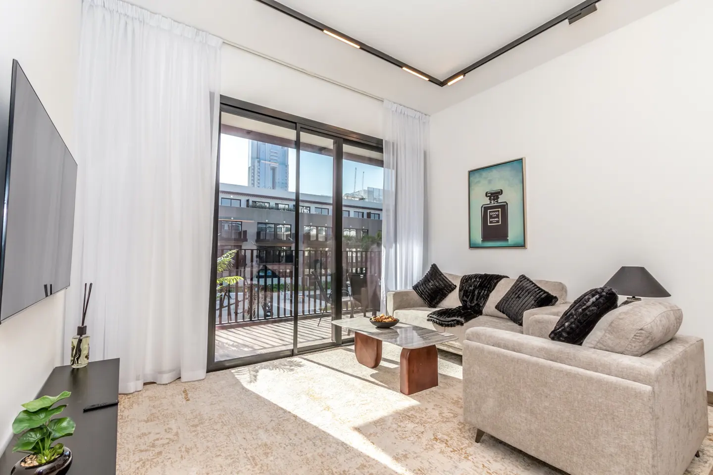 Bright living room with beige sofas, black pillows, and a marble coffee table. Balcony with city view visible through sliding glass doors.