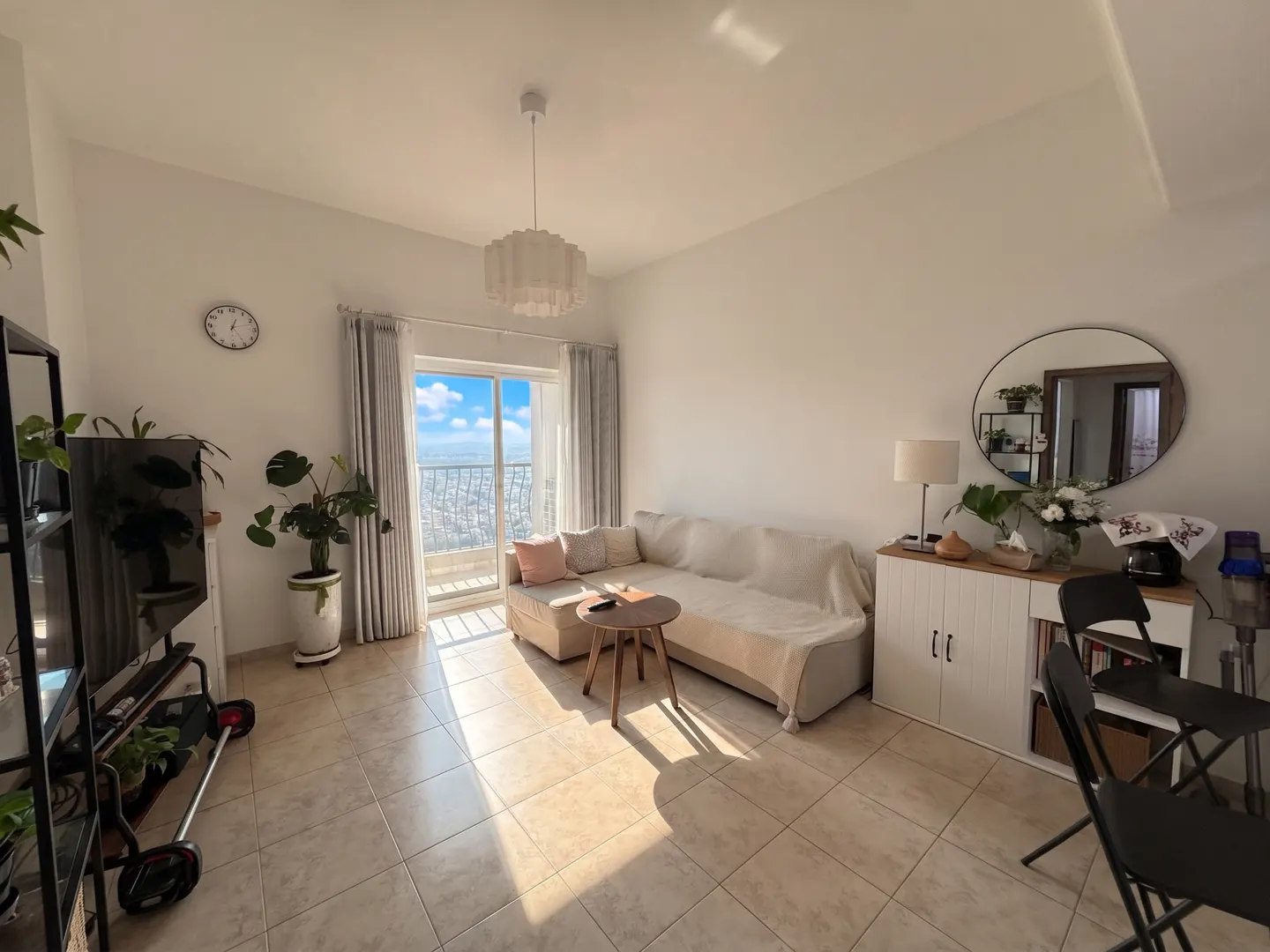 Bright living room with beige sofa, round table, and city view from balcony. Plants and a round mirror add to the decor.