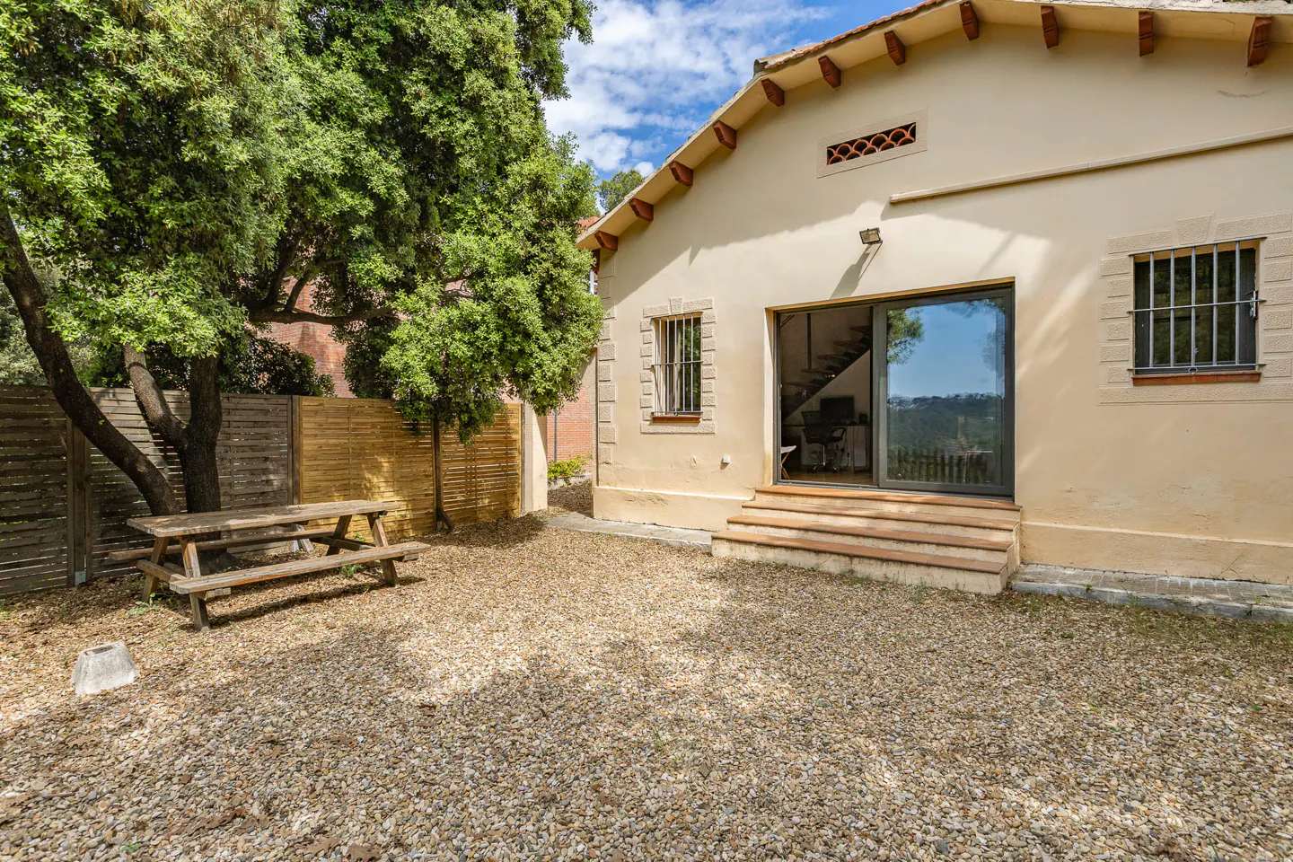Backyard view of a beige house with a picnic table, gravel ground, and a sliding glass door leading to stairs inside.