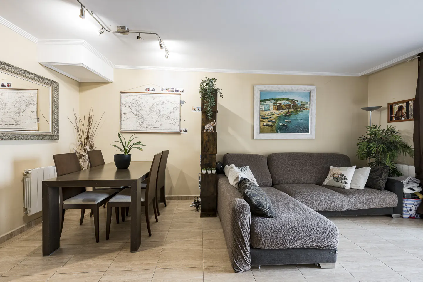A living and dining area with beige walls, a dark wood table and chairs, and a gray sectional sofa. Artwork and plants decorate the space.