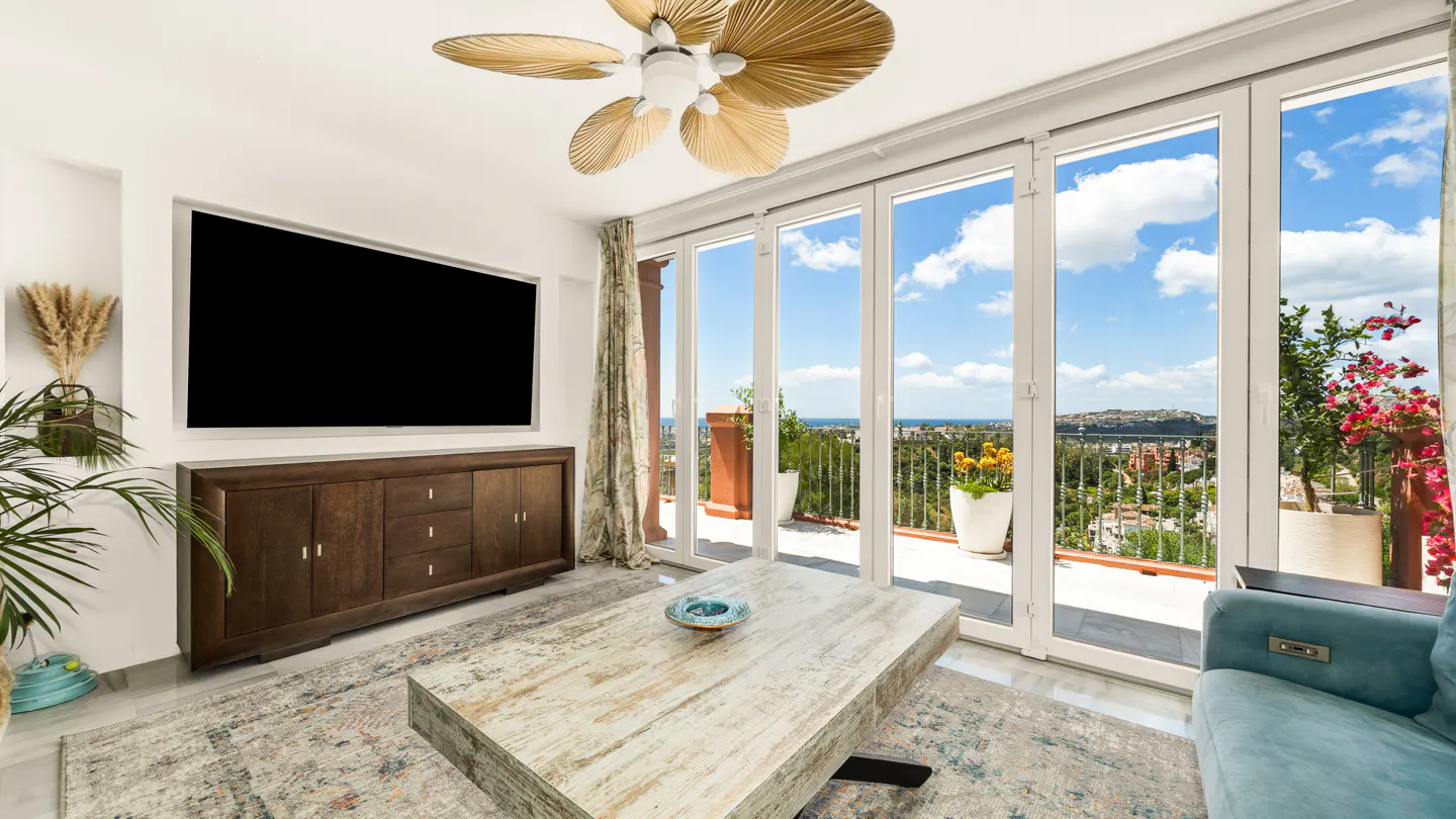 Bright living room with a large TV, wood cabinet, and a light wood coffee table. Floor-to-ceiling windows offer a view of a sunny landscape.
