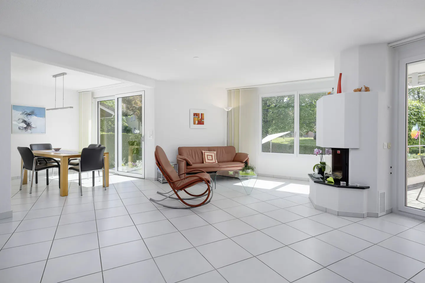 Bright, open-plan living room with white tile floor, dining table, brown sofa, rocking chair, and modern white fireplace.