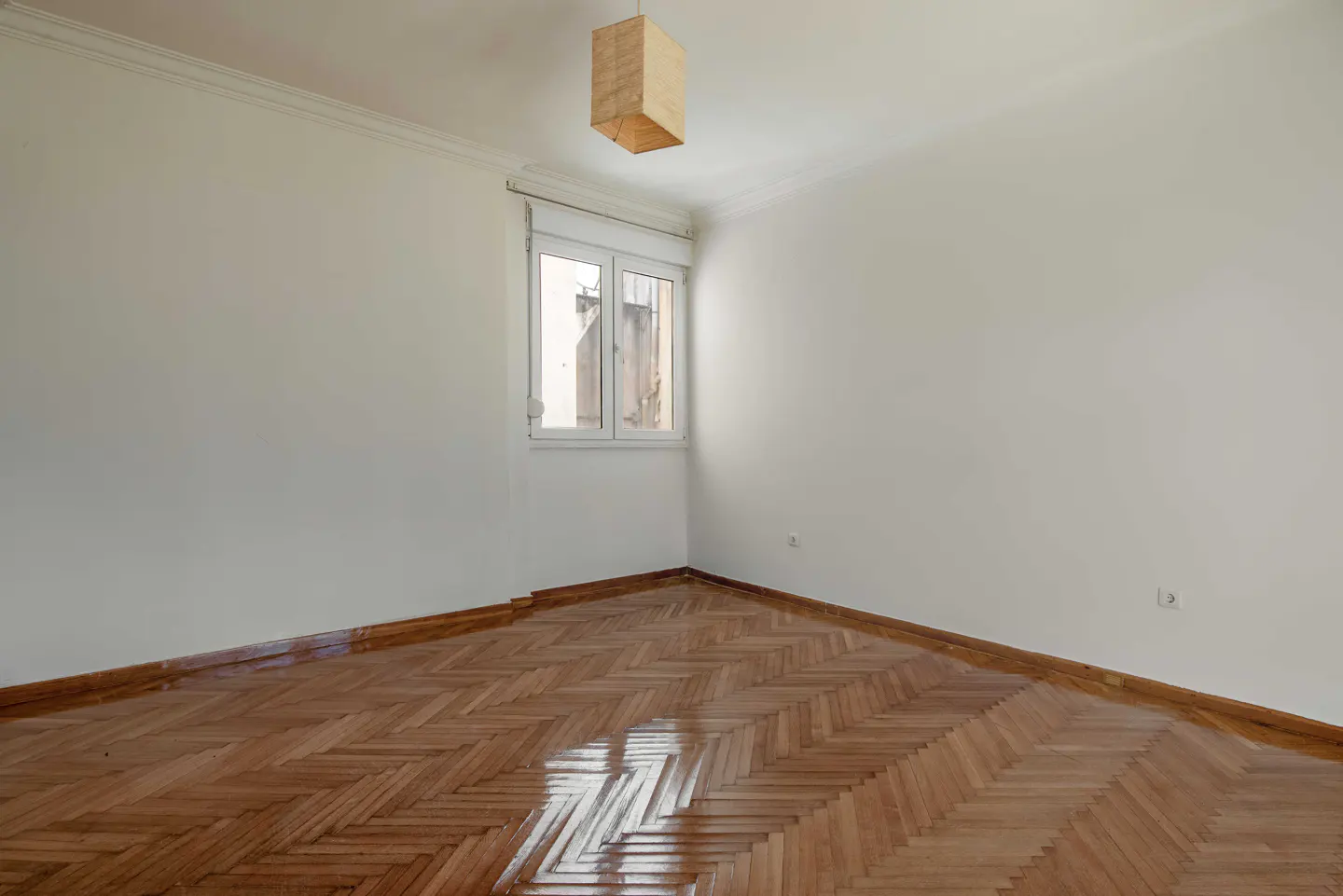 Bright, empty room with white walls, herringbone wood floor, and a window. A square, tan lampshade hangs from the ceiling.