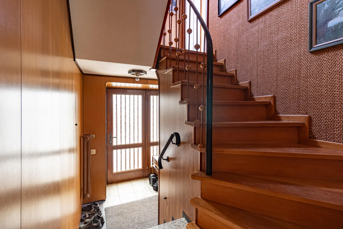 Interior view of a home's entryway with wooden stairs, a black metal railing, and a front door with vertical slats.
