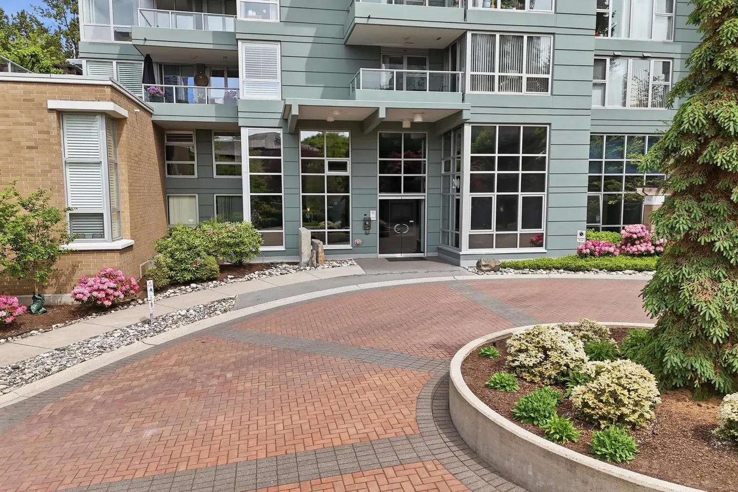 Exterior view of a modern, multi-story condo building with a brick driveway and landscaped entrance. The building is light green with large windows.
