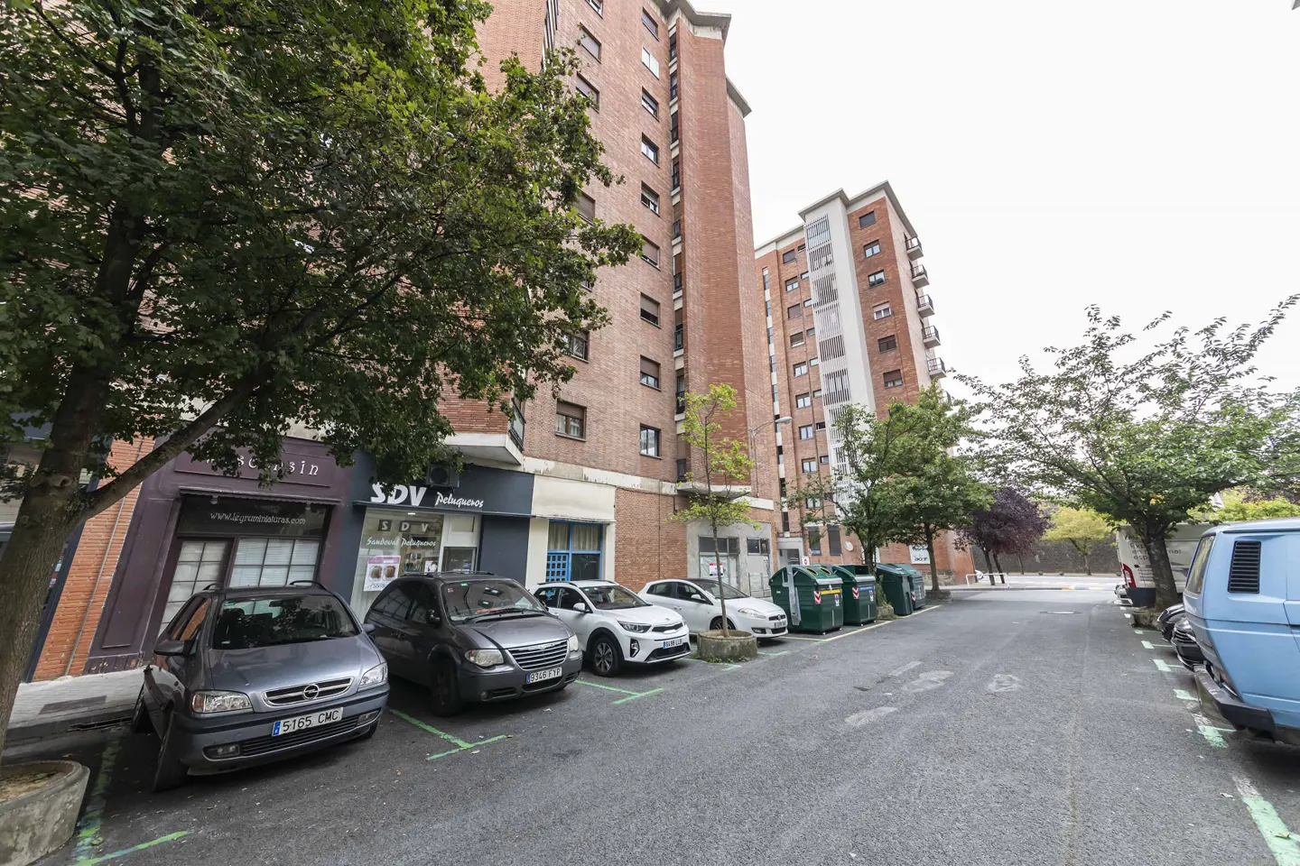 Street view of tall brick apartment buildings with parked cars and green recycling bins.