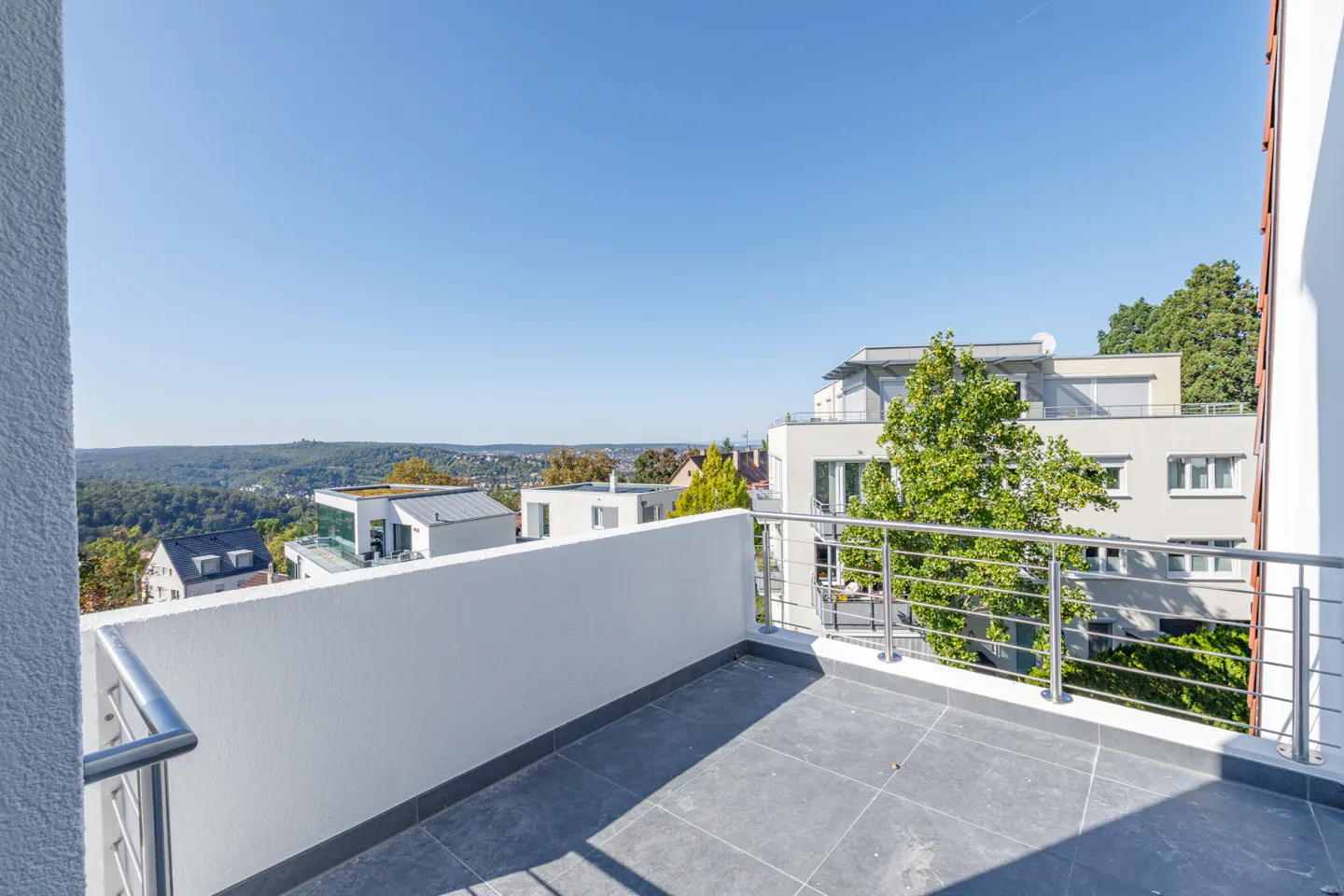 View from a gray tiled balcony with a white wall and metal railing, overlooking houses and trees under a clear blue sky.