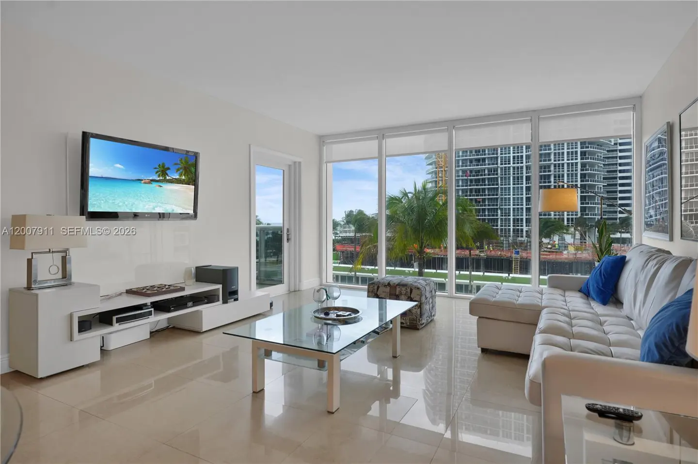 Bright living room with white sectional sofa, glass coffee table, and large windows overlooking city buildings and palm trees. A TV displays a beach scene.