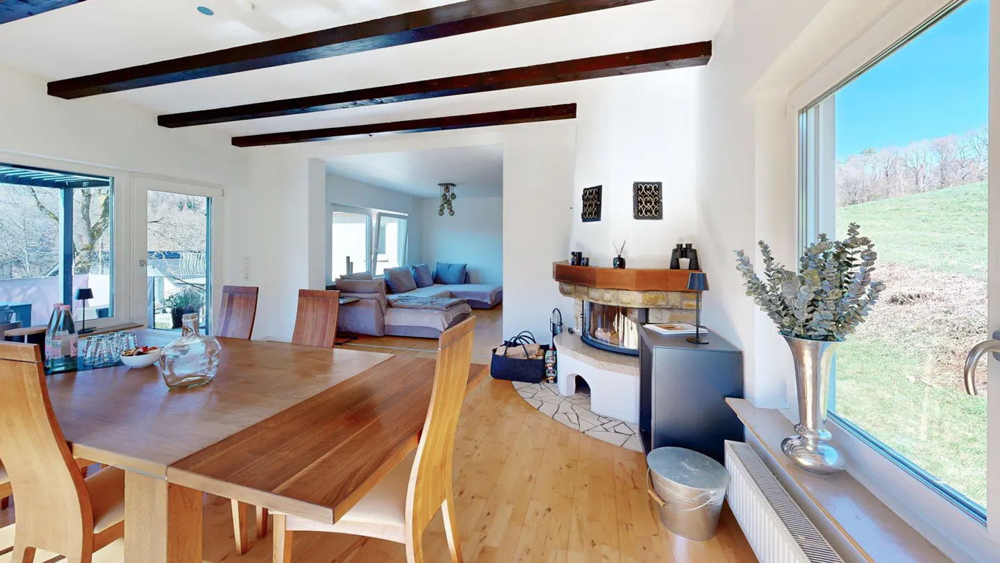 A bright, open-concept living space with a wooden dining table, fireplace, and large window with a view of a green hillside.