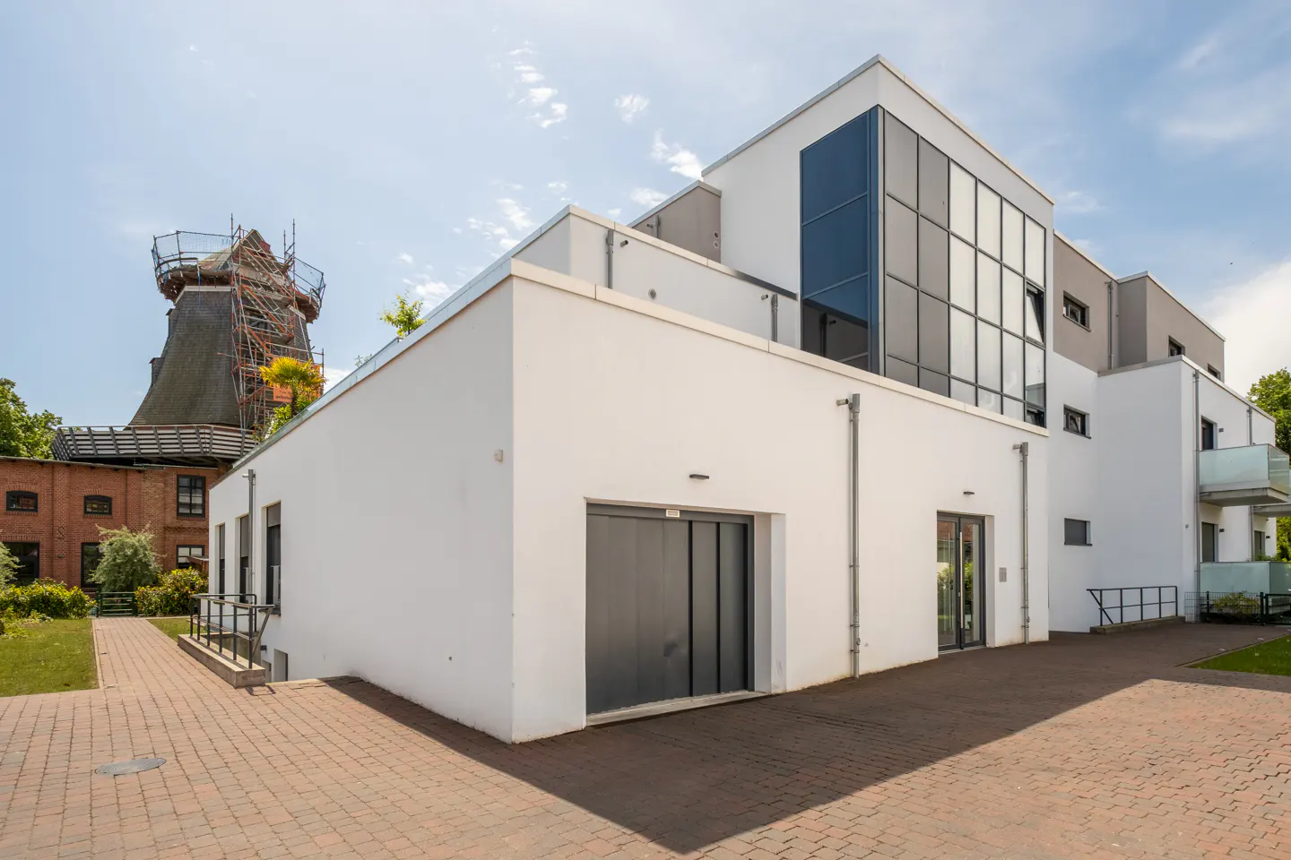 Modern white building with glass panels and a garage, set next to a historic brick windmill under a blue sky.