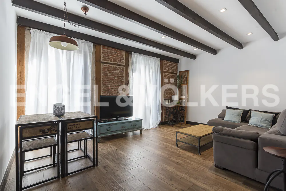 Living room with exposed brick, wood beams, and hardwood floors. A gray sofa and industrial-style table and chairs furnish the space.