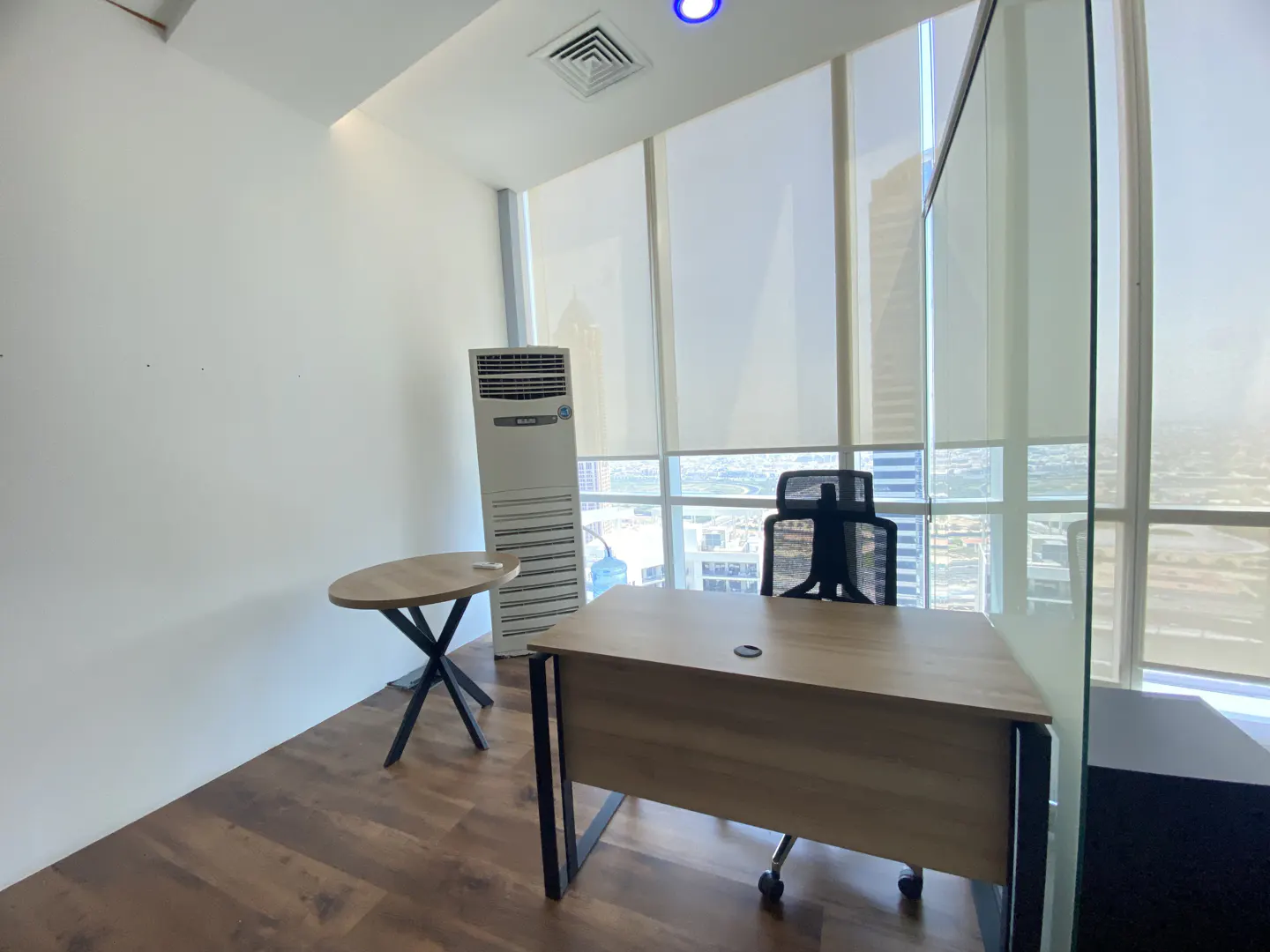 Office space with a desk, chair, and small table. Large windows offer a city view. The walls are white, and the floor is wood.