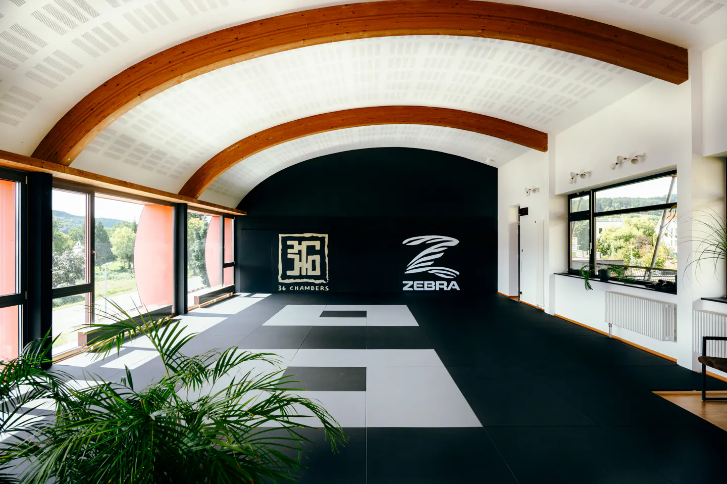 A bright martial arts studio with black and white mats, logos on the back wall, and large windows.