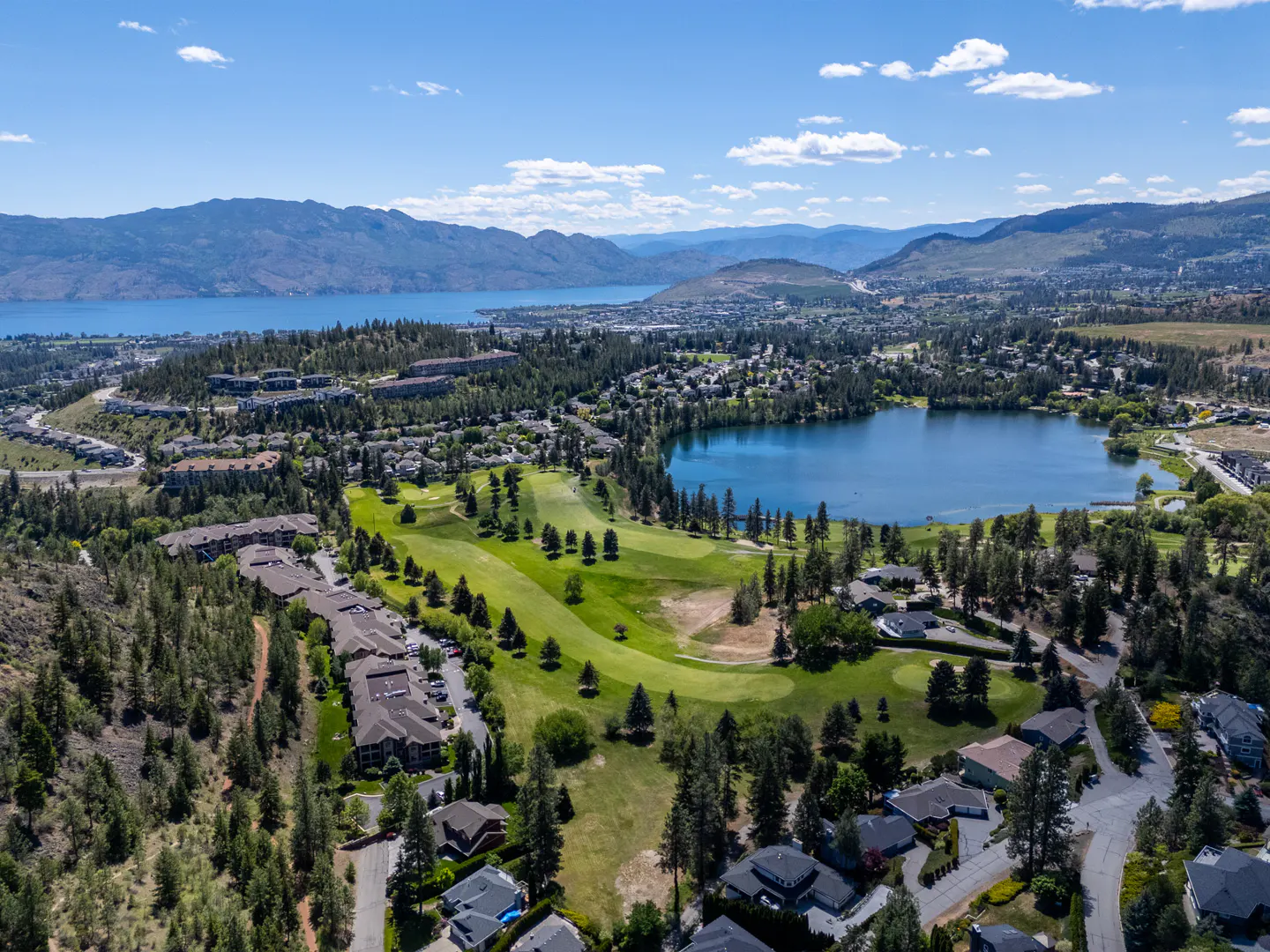 Aerial view of a green golf course, lake, and mountains under a blue sky with scattered clouds. Houses and trees surround the golf course.