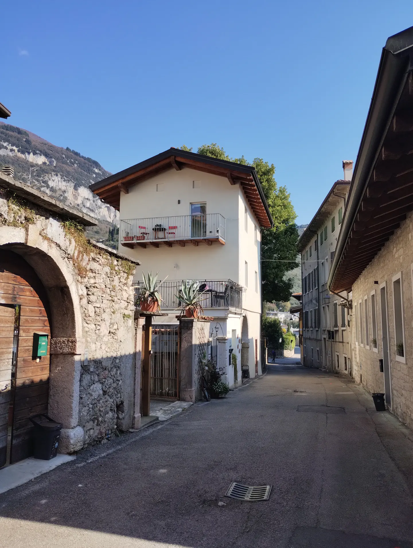 Street view of a two-story white house with balconies, stone archway, and mountain backdrop under a clear blue sky.