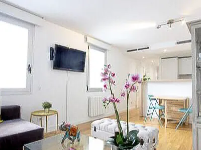 Bright, modern apartment interior with white walls, wood floors, and a TV. Pink orchids sit on a white ottoman in the foreground.