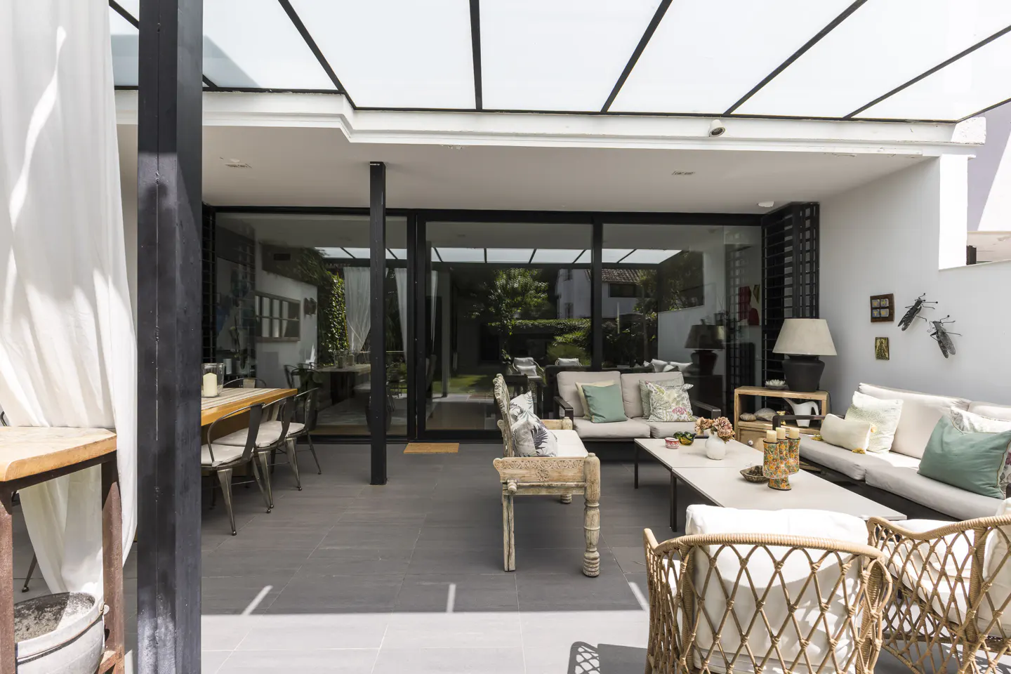 Bright sunroom with glass ceiling, gray tile floor, and white walls. Wicker chairs and a sofa with pillows provide seating. A dining table is visible through glass doors.