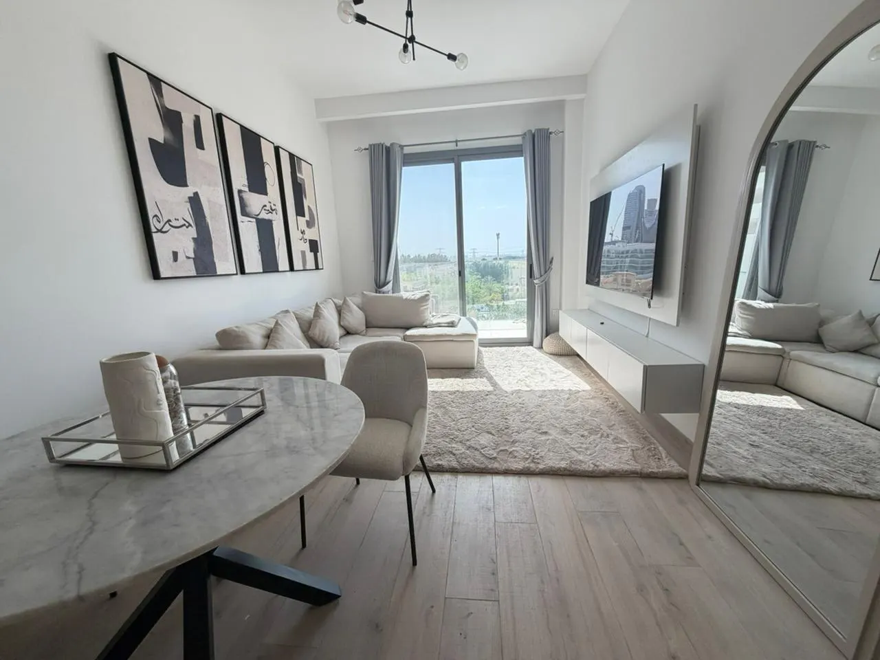 Bright living room with a marble table, beige sectional sofa, and a large window with a city view. Abstract art hangs on the wall.
