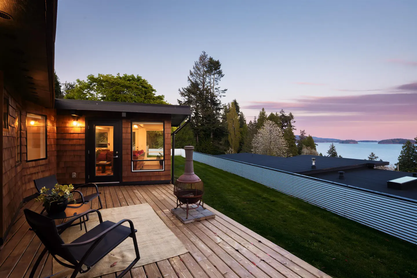 Wooden deck with chairs and a chiminea overlooks a grassy yard and ocean view at sunset. The house has cedar siding.